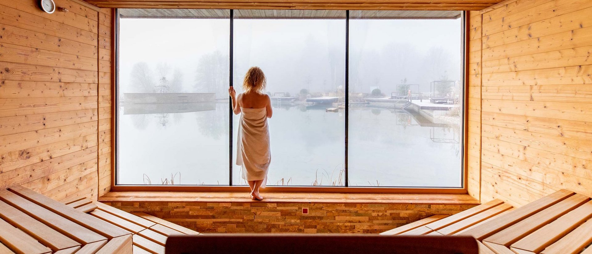 Person dans sauna regarde à travers une grande fenêtre un lac brumeux