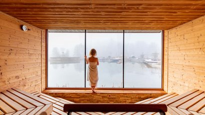 Person im Saunaraum blickt durch großes Fenster auf nebligen See