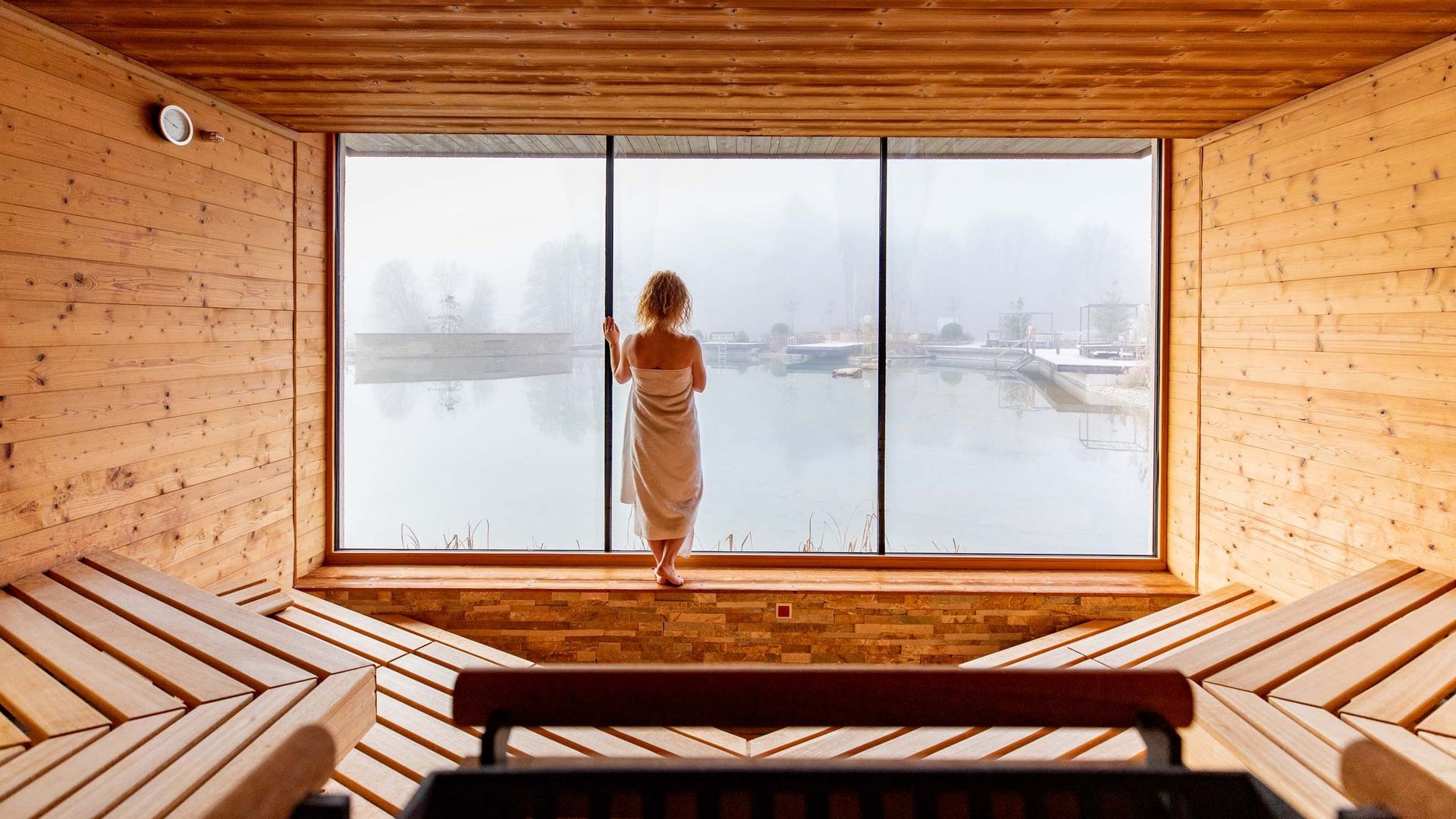 Person in sauna room looking out large window at misty lake