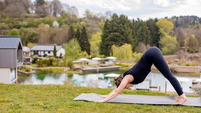 Frau macht Yoga-Pose auf Matte draußen mit See und Bäumen im Hintergrund