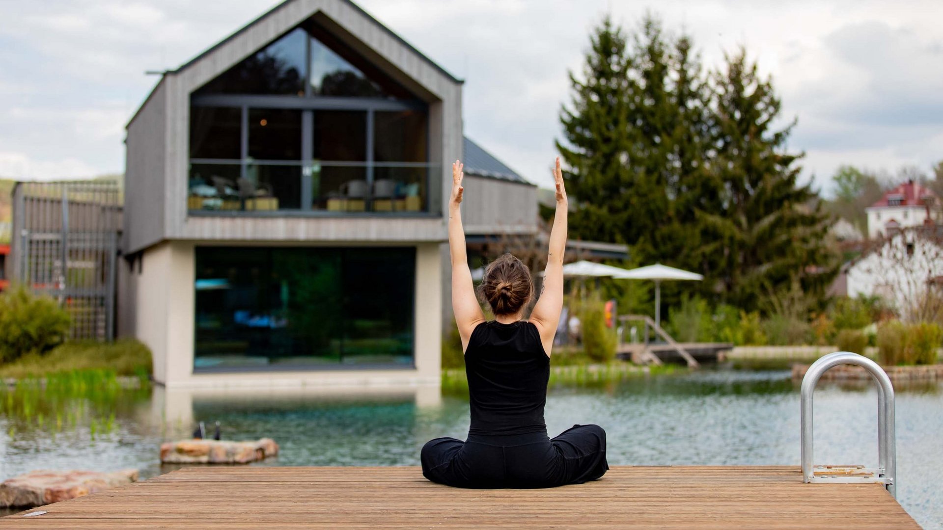 Frau macht Yoga auf Holzsteg vor modernem Haus an einem Wasserteich