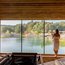 Frau in Sauna blickt auf See und Wald durch großes Fenster