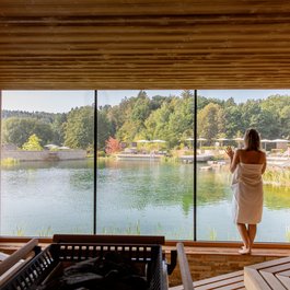 Frau in Sauna blickt auf See und Wald durch großes Fenster