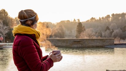 Frau in Winterjacke schaut auf einen Teich und hält dampfende Tasse
