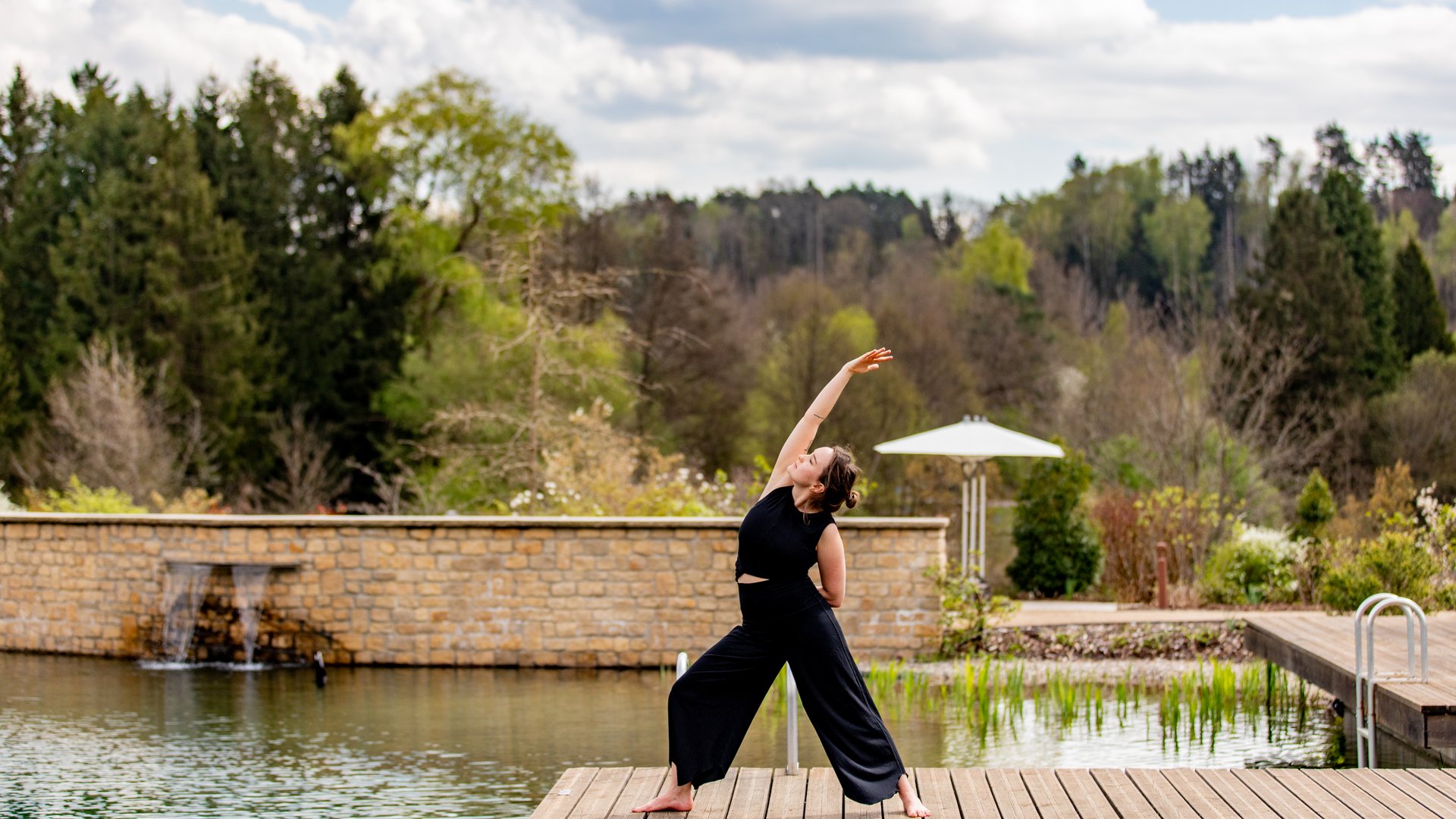 Frau macht Yoga auf Holzsteg neben Teich und Garten