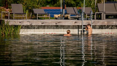 Zwei Kinder schwimmen in einem natürlichen Pool neben Gartenstühlen