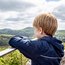 Junge in blauer Jacke blickt von einer Aussichtsplattform auf bewaldete Hügel