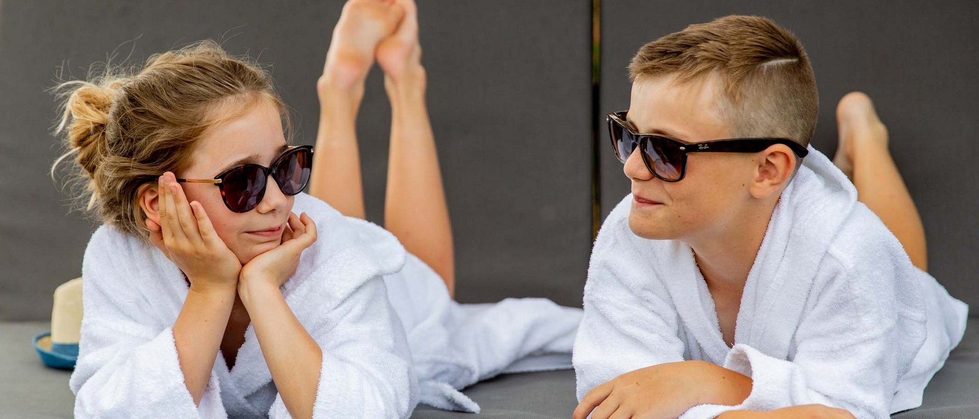 Zwei Kinder liegen in Bademänteln und Sonnenbrillen auf Liegen draußen