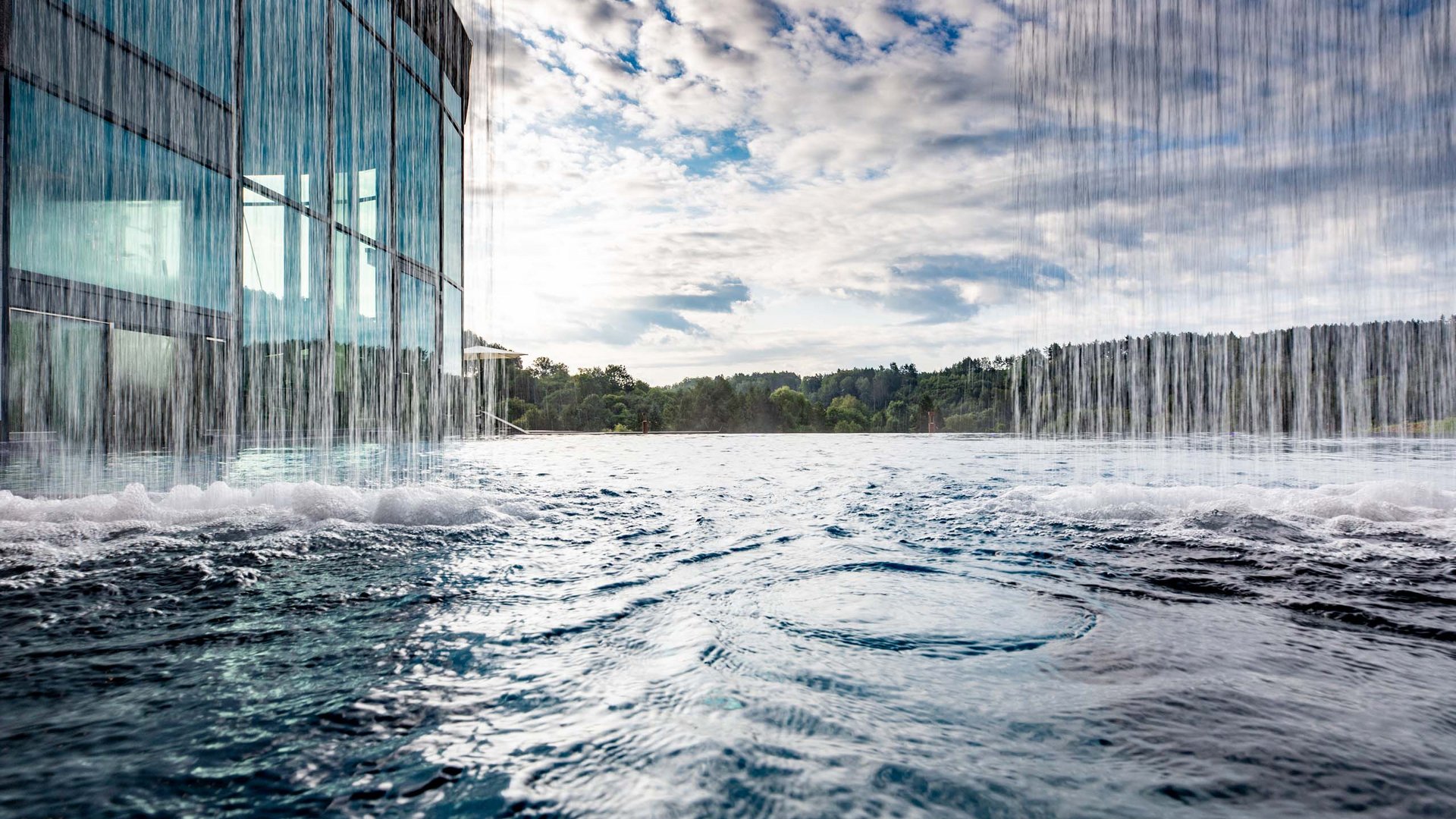Cascade dans une piscine à débordement avec façade en verre moderne et ciel nuageux