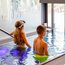 Zwei Kinder mit Badesachen stehen im Hallenbad mit Glasfenstern