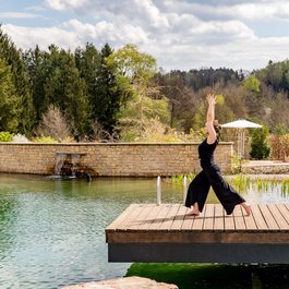 Frau macht Yoga auf Holzsteg am See mit Bäumen im Hintergrund