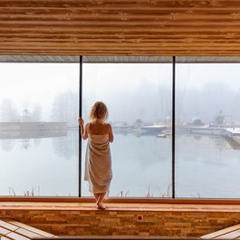 Frau im Handtuch blickt aus einem holzverkleideten Raum auf ein nebliges Wasserbecken