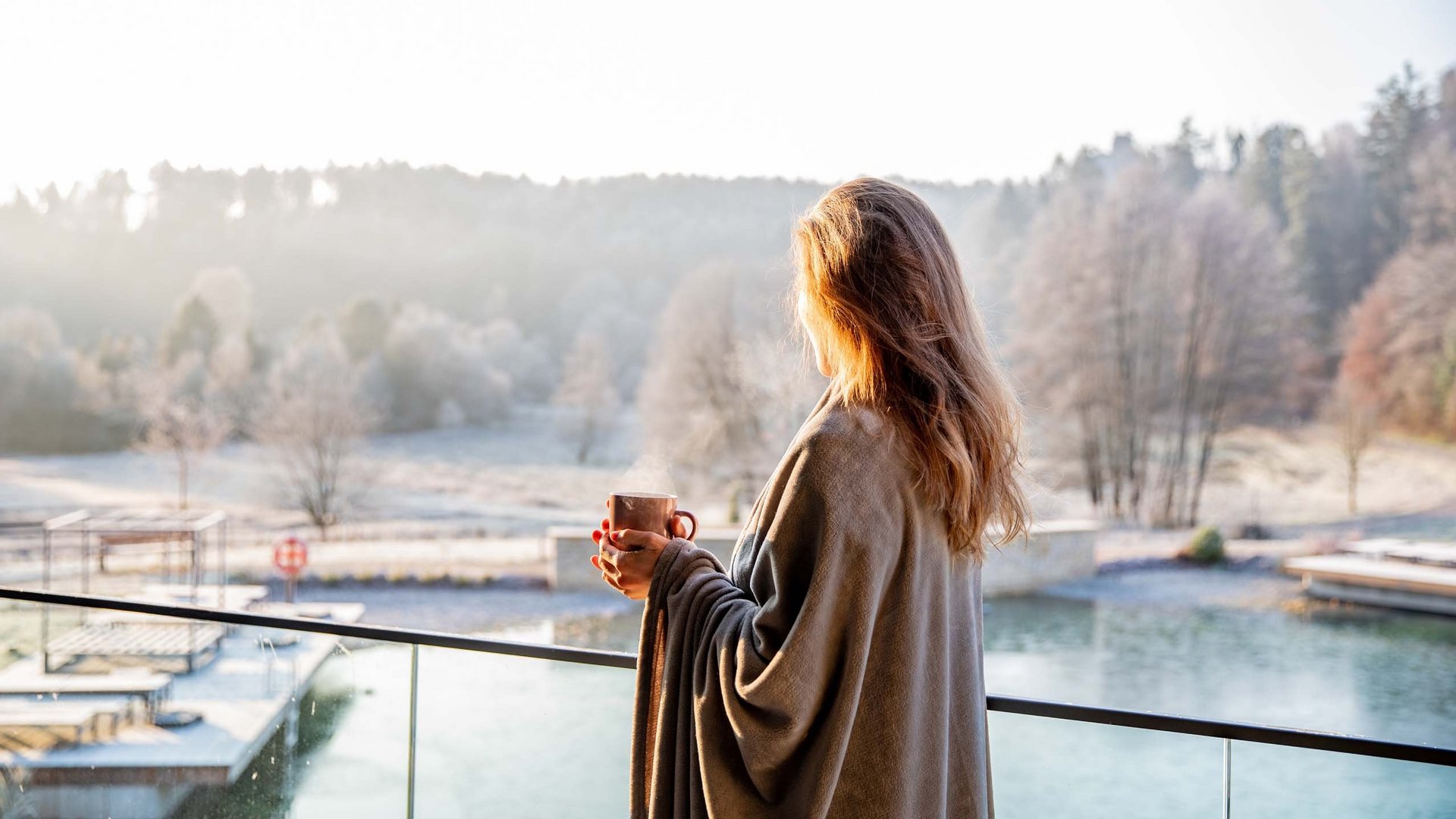 Frau mit Tasse auf Balkon blickt auf winterliche Landschaft mit See